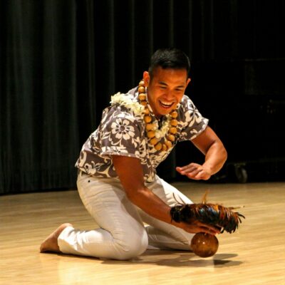 John-Mario, wearing white pants and a gray, black and white flowered print aloha shirt, is kneeling on the floor dancing "Heʻeia," a hula noho (seated hula) with an ʻuliʻuli (feathered gourd rattle that is used as a hula implement).